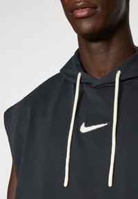 Hoodie senza maniche Nike in tessuto grigio scuro, con logo bianco e lacci color crema. Texture liscia e vestibilità rilassata.