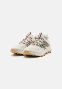 Zapatillas deportivas ligeras de color beige con una parte superior texturizada y suela de goma. Presentan acentos en verde olivo contrastantes y elementos de diseño aerodinámico.