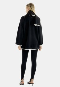 Pullover nero oversize con borchie bianche, dotato di un alto colletto e maniche lunghe, indossato sopra leggings neri e sandali con tacco.