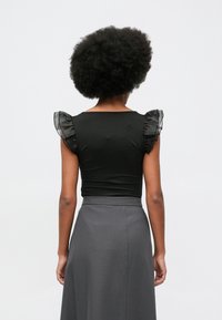 Kobieta z kręconym afro widziana od tyłu, ubrana w czarną bluzkę z przezroczystymi, falbaniastymi krótkimi rękawami oraz spódnicę w kolorze szarym z wysokim stanem.