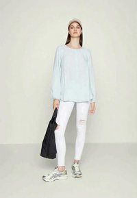 Camisa de manga longa azul clara com decote redondo, jeans skinny brancos rasgados, casaco preto e sapatos de desporto prateados. Boné minimalista.