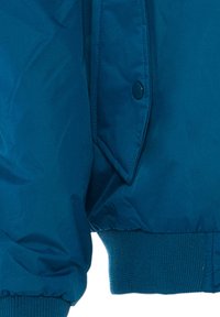 Giacca a vento teal con un esterno liscio e lucido, polsini a costine e chiusura a scatto. Il tessuto è leggero e resistente.