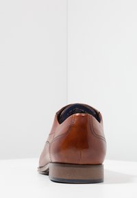 Chaussure habillée en cuir brun avec une texture lisse, un talon arrondi et des détails de couture subtils. La vue arrière met en valeur le design épuré et le savoir-faire.