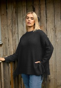 Czarny sweter dzianinowy o prążkowanej teksturze, z bocznymi kieszeniami i frędzlami. Zestawiony z jasnoniebieskimi dżinsami. Naturalne drewniane tło.