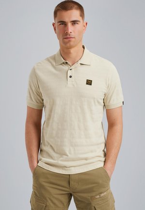 Camisa polo de manga corta color beige con patrón texturizado, tapeta de tres botones y un pequeño parche de logo negro en el pecho.