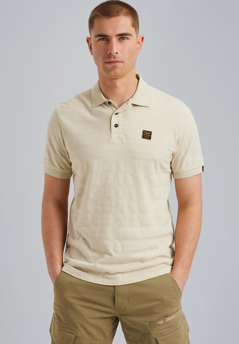 Kortærmet beige polo med et tekstureret mønster, tre-knaps lukning og et lille sort logo patch på brystet.