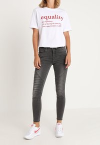 Camiseta blanca con texto rojo que dice "igualdad", combinada con jeans skinny grises con detalles acanalados, llevada con zapatillas blancas con detalles rosas.