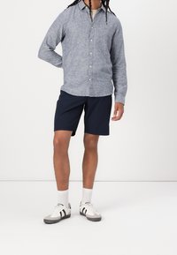Grijze overhemd met een gestructureerde stof, gecombineerd met marineblauwe shorts. Schoenen zijn wit met zwarte strepen, aangevuld met witte sokken.