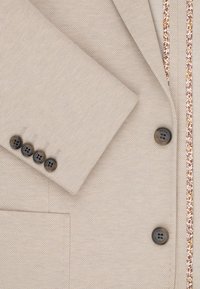 Beżowa żakiet o fakturze z klapami, kieszenią na piersi i pięcioma ciemnymi guzikami, z wzorzystym wykończeniem wzdłuż krawędzi.