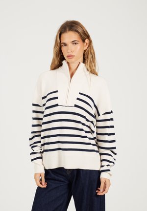 Jonge vrouw in een crèmekleurige en marineblauwe gestreepte half-rits trui en donkerblauwe jeans tegen een effen achtergrond.