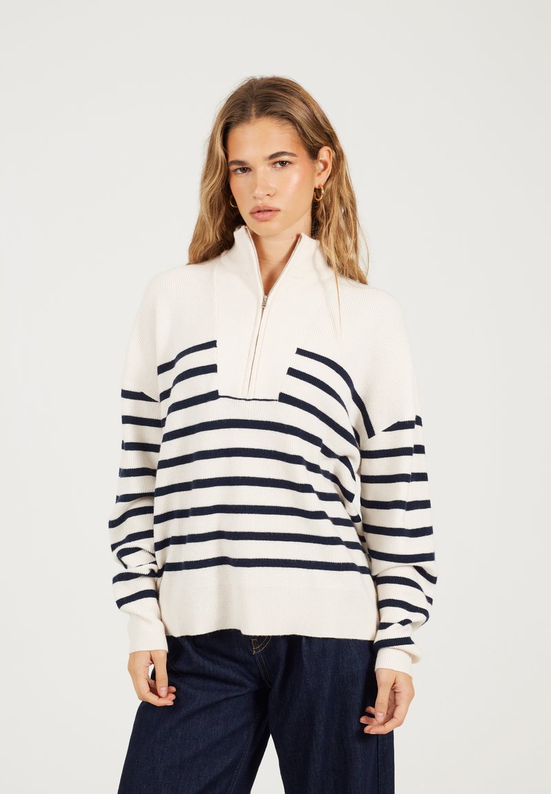 Młoda kobieta ubrana w sweter z kremowo-granatowymi paskami z półzamkiem i ciemnoniebieskie dżinsy na jednolitym tle.