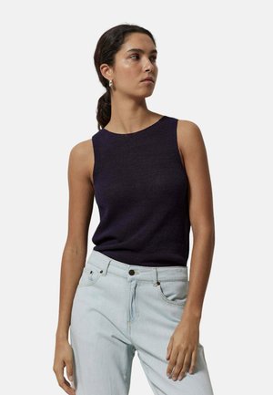 Mujer con cabello oscuro recogido, que lleva una camiseta negra sin mangas y vaqueros azul claro, de pie con expresión neutra y mirando hacia un lado.