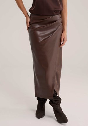 Femme portant une jupe midi en cuir marron avec un détail de ceinture asymétrique et des bottes molles en daim marron foncé.