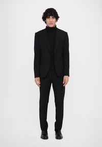 Schwarzes Anzugensemble, bestehend aus einem taillierten Blazer und passender Hose, getragen über einem schwarzen Rollkragenpullover. Glattes Material, maßgeschneiderter Schnitt.