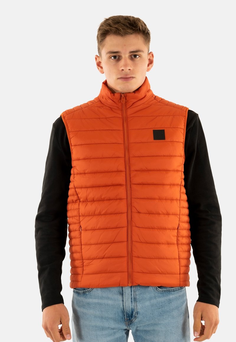 Oranje gewatteerde vest met horizontale stiksels en hoge kraag, voorzien van een rits aan de voorkant en een klein zwart logo-lable. G gedragen over een zwart shirt.
