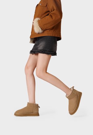 Persona che cammina indossando stivaletti color cuoio, gonna di jeans nera sfrangiata e giacca marrone con polsini in pelliccia su sfondo chiaro.