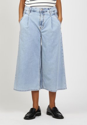 Person trägt hellblaue, weit geschnittene, gekürzte Jeanshosen, schwarze Loafers und ein weiß-navy gestreiftes Oberteil, Hände in den Taschen.