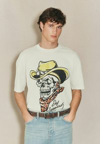 Kreem värvi t-särk, millel on graafika skeletist cowboy mütsiga ja mustripärase bandanaga. Kombineeritud helesiniste teksade ja pruuni vööga.