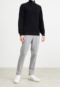 Personne portant un pull noir à col zippé à manches longues, un pantalon gris clair et des baskets blanches, debout sur un sol clair avec un fond uni.