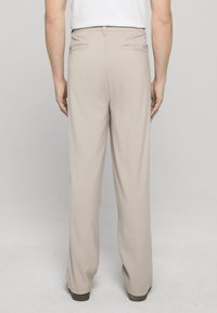 Pantaloni beige con una texture liscia, gambe dritte e tasche posteriori. La cintura presenta passanti per cintura. Indossati con una camicia bianca.