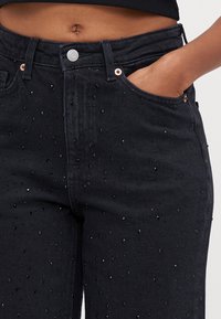 Jeans de denim preto com um design ajustado e cintura média, adornados com pequenas contas pretas para textura e detalhe.