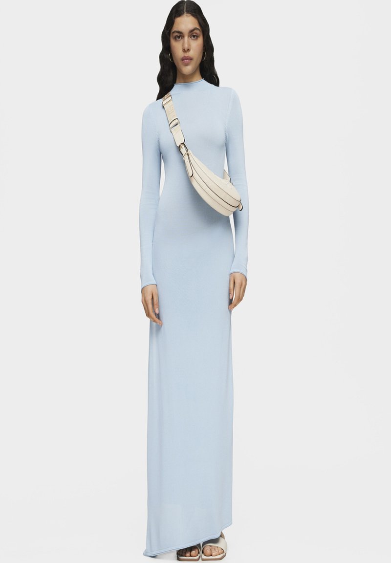 Vestido maxi de manga larga en un diseño ajustado de color azul claro. Acompañado de un bolso cruzado de color crema con forma curva y correa ajustable.