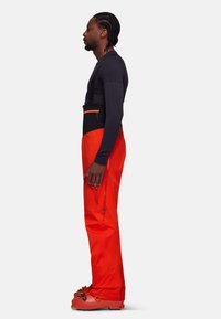 Des pantalons de ski rouges présentent des bretelles ajustables, une section de taille noire et des poches zippées. Portés avec des bottes de ski orange assorties.