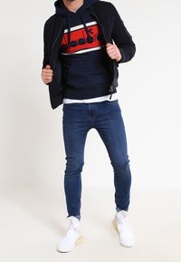 Veste bomber marine sur un sweat à capuche rouge et blanc à rayures, associée à un jean skinny bleu et des baskets blanches. Tenue urbaine décontractée.