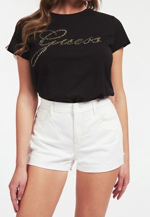 Nainen, jolla on musta lyhythihainen paita, jossa strassikirjaimia, ja valkoiset korkeavyötäröiset farkkushortsit yksiväristä taustaa vasten.