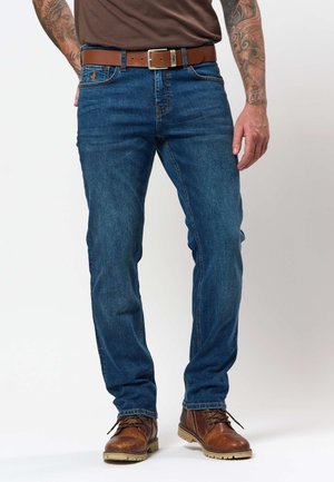 Férfi, aki kék farmert, barna bőr övet, barna csizmát és sötétbarna inget visel, tetovált karok láthatók.