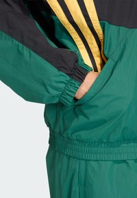 Grön och svart vindjacka med gula ränder, utrustad med elastiska muddar och midjeband, lätt material och sidofickor.