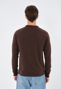 Cămașă lungă, maro, cu mâneci raglan și guler rotund, purtată cu blugi din denim deschis la culoare, prezentată din spate.