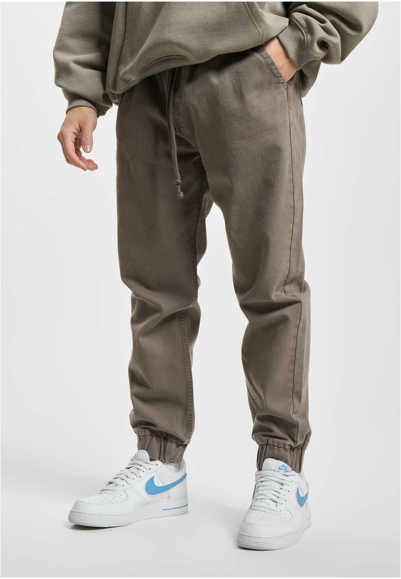 Personne portant un pantalon de jogging gris et des baskets blanches avec un swoosh Nike bleu clair, debout avec une main dans la poche.