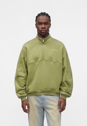 Ung mand med flettet hår iført en olivengrøn Jordan sweatshirts med kvart lynlås og lyseblå vaskede jeans, stående mod en ensfarvet baggrund.