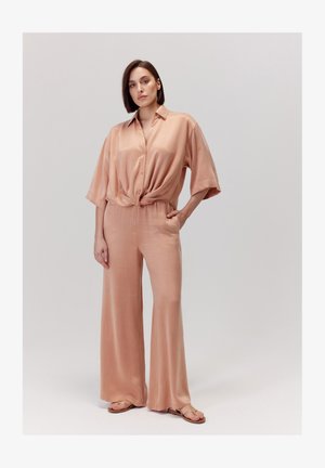 Brzoskwiniowy satynowy kombinezon z węzłem z przodu, krótkimi rękawami i szerokimi spodniami. Płaskie sandały dopełniają stylizację.