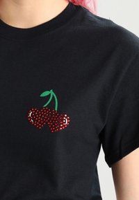 Svart bomullströja med en rhinestone-körsbärdesign i rött med gröna löv, placerad på vänster bröst.