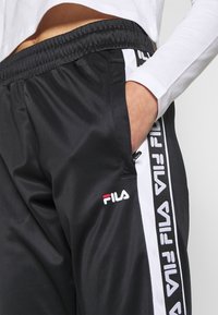 Černé sportovní kalhoty s elastickým pasem, bílými postranními pruhy a logem Fila. Materiál je hladký a mírně lesklý, s neformálním designem.