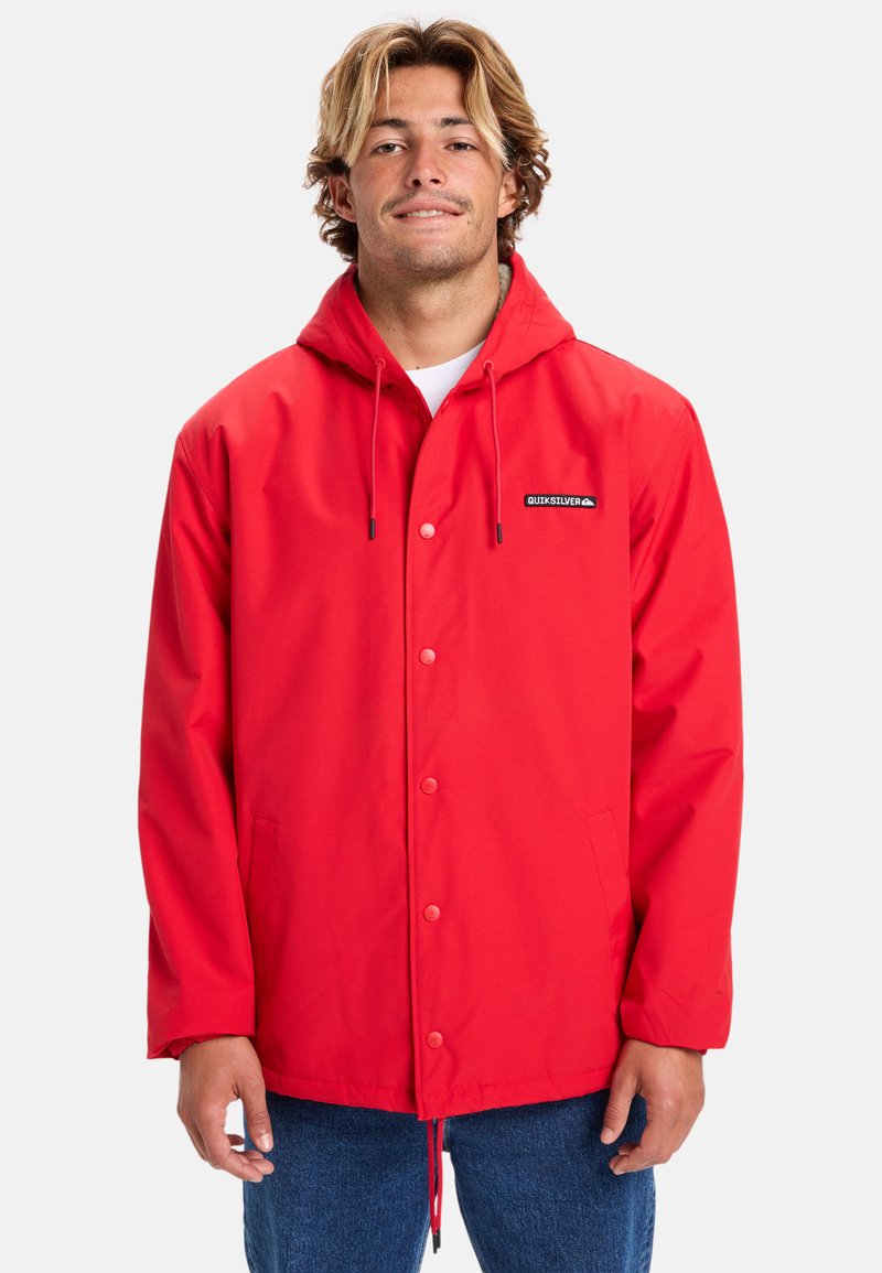 Rød vandtæt jakke med hætte, frontlukning med trykknapper, sidelommer og et logo-patch på brystet. Glat tekstur, casual pasform.