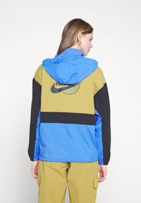 Nike vindjakke i blå, khaki og svart. Har hette, elastiske mansjetter og en stor logo trykt på baksiden. Teksturert materiale.