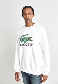 Fehér pulóver zöld krokodil logóval és "LACOSTE" felirattal. Puha pamutból készült, kerek nyakkivágással és bordázott mandzsettákkal.