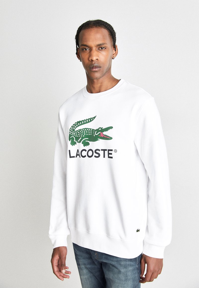 Fehér pulóver zöld krokodil logóval és "LACOSTE" felirattal. Puha pamutból készült, kerek nyakkivágással és bordázott mandzsettákkal.