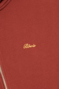 Rustfarget stoff med en liten gul brodert "Bkvis"-logo nær en sølvglidelås på venstre side.