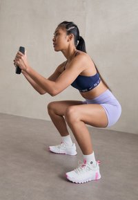 Nike sport BH i marineblå, højtaljede lilla shorts, hvide sneakers med lyserøde detaljer. Udføreren squatter mens hun holder en sort enhed.