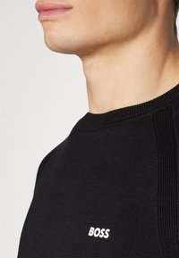 Schwarzer Strickpullover mit geripptem Kragen und seitlichen Akzenten, mit einem weißen "BOSS"-Logo auf der Vorderseite. Nahaufnahme, die sich auf den Kragen konzentriert.