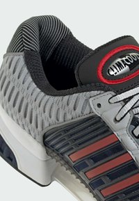 Grijze sportschoen met een bovenwerk van getextureerd mesh, zwarte accenten, rode strepen en een reflecterend logo; voorzien van een gewatteerde kraag en een rubberen zool.