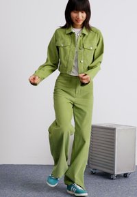 Groene corduroy jack en bijpassende brede broek, gecombineerd met een witte top en teal sneakers, op een effen achtergrond.