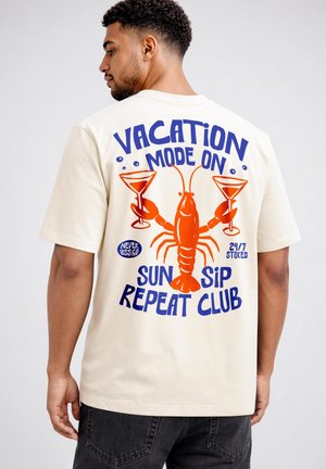 Man wearing beige t-shirt with a red lobster holding cocktail glasses and text "Vacation Mode On, Sun Sip Repeat Club" on the back.
