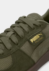 Gros plan d'une basket Puma Palermo vert olive avec des lacets en tissu et une étiquette logo dorée sur le panneau latéral texturé.