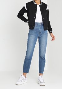 Zwarte varsityjas met witte strepen, gedragen over een wit T-shirt. Vervaagde blauwe straight-leg jeans en witte sneakers met grijze accenten.