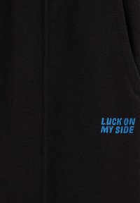 Črn material z gladko teksturo; vključuje modro vezeno besedilo "LUCK ON MY SIDE." Oblikovanje je preprosto, brez dodatnih vzorcev ali poudarkov.